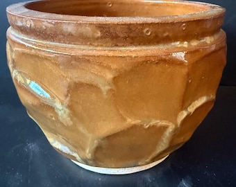 Pode incluir: Um vaso de cerâmica redondo com um esmalte brilhante castanho-mel e uma textura exterior geométrica facetada. O rebordo apresenta um padrão de pontos subtil e repetitivo, e a base tem um acabamento com um rebordo circular branco não vidrado.
