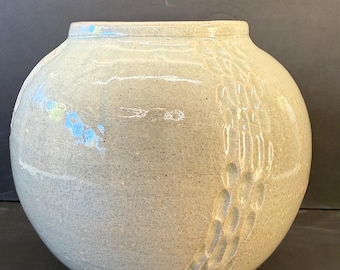 Pode incluir: Um vaso de cerâmica esférico com um esmalte brilhante cinza claro salpicado. A superfície apresenta uma faixa vertical de impressões ovais texturizadas e reentradas no lado direito. O vaso tem um pescoço curto e estreito e um corpo largo.