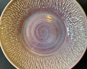 Pode incluir: Um prato redondo de cerâmica com um centro liso em espiral em tons de roxo e azul, rodeado por uma borda larga e texturizada com um padrão repetitivo tipo favo de mel em bege claro com reflexos sutis de esmalte roxo.