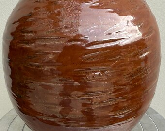 Pode incluir: Um vaso grande e redondo de cerâmica vidrada em tons de castanho. A superfície tem um padrão texturizado e estriado. O vaso está sobre uma superfície metálica prateada. A forma geral é esférica, com uma base ligeiramente achatada.