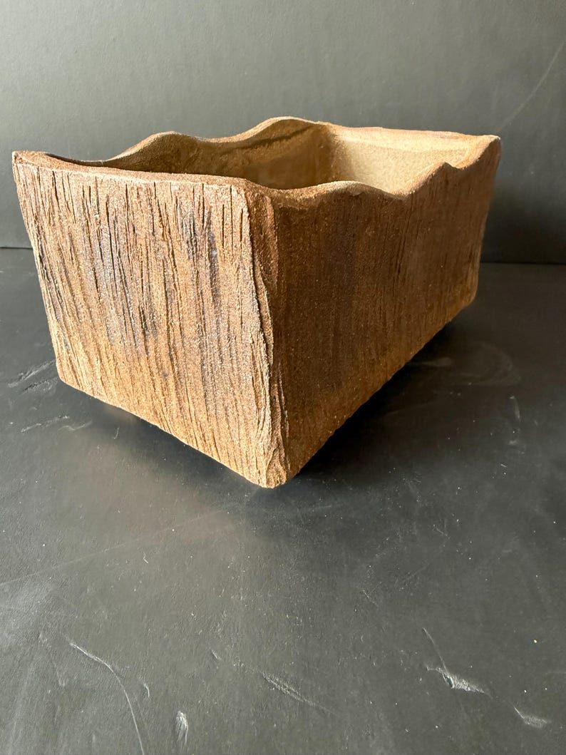 Pu&ograve; includere: Un vaso rettangolare in ceramica con un esterno marrone ruvido e testurizzato che imita l'aspetto della corteccia d'albero. Il contenitore presenta un bordo superiore ondulato e irregolare e un interno cavo, con una finitura naturale e terrosa.