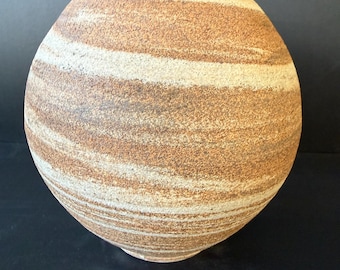 Pode incluir: Um vaso de cerâmica esférico com uma superfície texturizada e mate, apresentando faixas horizontais e onduladas em tons terrosos de castanho, bege e creme. O vaso tem uma borda preta lisa na abertura, contrastando com o corpo áspero e arenoso.