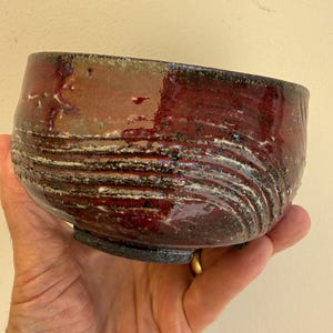 May include: A ceramic bowl with a deep red and dark brown glaze, featuring a textured surface with horizontal grooved lines. The bowl has a wide, rounded shape with a dark, rough-textured base and a glossy finish that reflects light.