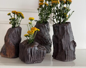 Può includere: Quattro vasi in ceramica marrone scuro con una superficie ruvida che ricorda la corteccia. Ogni vaso contiene un piccolo mazzo di fiori giallo brillante con steli verdi. I vasi variano in altezza e forma, richiamando tronchi d'albero.