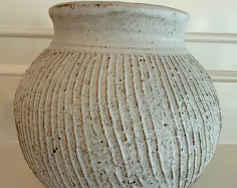 Pode incluir: Um vaso de cerâmica branco sujo com uma superfície texturizada e formato arredondado. O vaso tem uma borda alargada e é decorado com linhas verticais esculpidas. Um elemento decorativo para a casa.