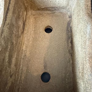 Pu&ograve; includere: Un vaso rettangolare con una superficie ruvida e maculata nei toni del beige e del marrone. La base interna presenta due fori di drenaggio circolari centrati lungo la lunghezza del contenitore, con una tonalit&agrave; terrosa uniforme.