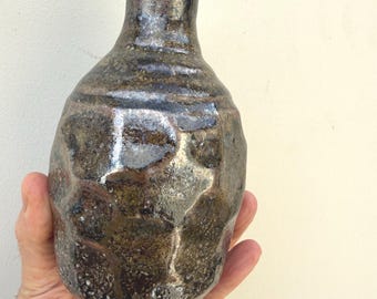 Pode incluir: Um vaso de cerâmica artesanal, com superfície texturizada e esmalte escuro e manchado. O vaso tem um pescoço estreito e um corpo arredondado. As cores incluem tons de castanho e preto, com reflexos metálicos.