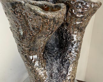 Pode incluir: Um vaso de cerâmica alto moldado à mão com uma superfície altamente texturizada e craterizada. O esmalte apresenta uma mistura complexa de tons castanho-escuro, carvão e bronze metálico. A borda é irregular e ondulada, criando uma abertura.