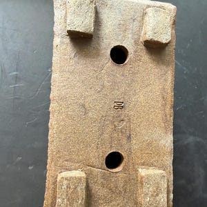 Pu&ograve; includere: Una base rettangolare per vaso in ceramica marrone con quattro piedini rialzati agli angoli, due fori di drenaggio circolari al centro e le lettere DB impresse sulla superficie ruvida tra i fori di drenaggio.