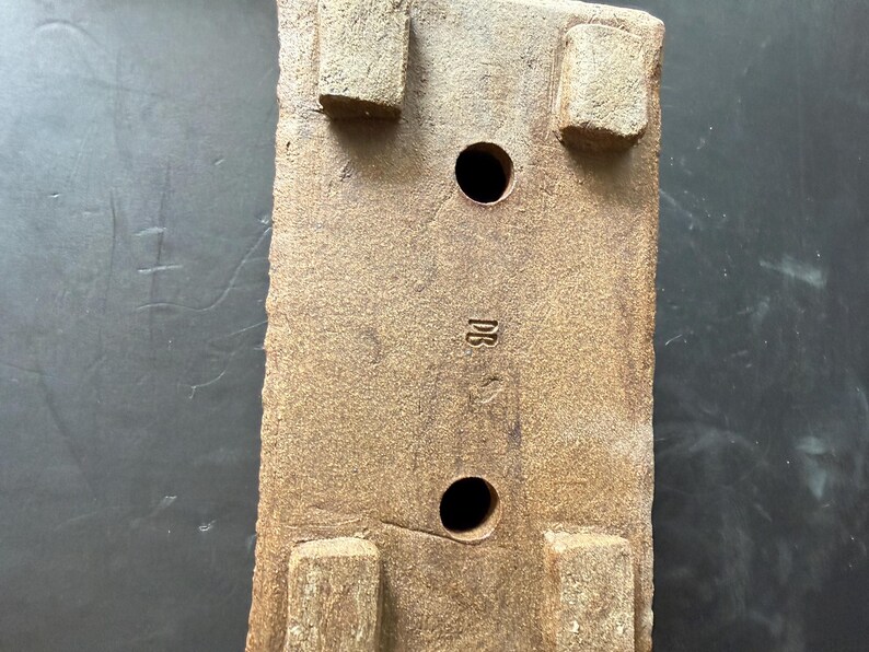 Pu&ograve; includere: Una base rettangolare per vaso in ceramica marrone con quattro piedini rialzati agli angoli, due fori di drenaggio circolari al centro e le lettere DB impresse sulla superficie ruvida tra i fori di drenaggio.