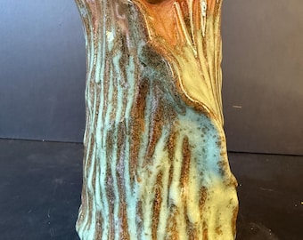 Pode incluir: Um vaso de cerâmica com uma superfície texturizada e orgânica que lembra a casca de uma árvore. O exterior apresenta uma mistura de tons castanhos terrosos e um esmalte verde-menta pálido que escorre pelas cristas verticais do corpo cilíndrico.