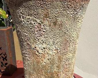 Può includere: Un vaso in ceramica alto e conico con una superficie molto strutturata e porosa. L'esterno presenta una tavolozza di colori screziati nei toni del beige terroso, verde tenue e rosa delicato, con un bordo superiore liscio e arrotondato.
