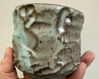 Pode incluir: Uma chávena de cerâmica feita à mão com uma superfície irregular e texturizada. O vidrado é uma mistura salpicada de tons verde-sálvia e castanho-terra, com pontos escuros. A base é de argila castanha escura não vidrada.