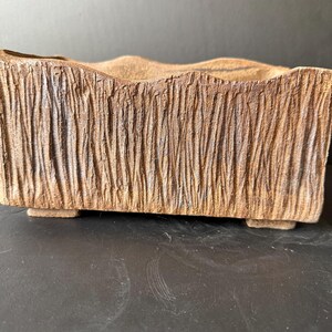 Pu&ograve; includere: Un vaso rettangolare in ceramica con un esterno marrone testurizzato che ricorda la corteccia. Il bordo superiore presenta un profilo ondulato e la base &egrave; sostenuta da quattro piccoli piedini bassi, conferendo un aspetto solido.