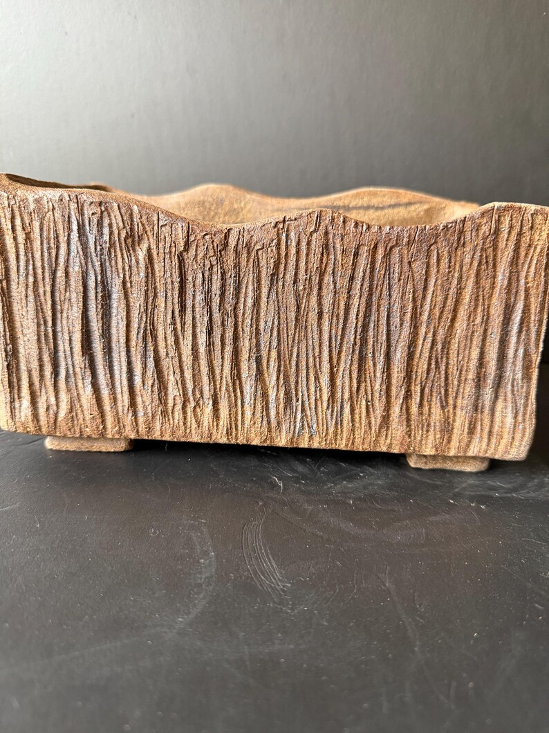 Pu&ograve; includere: Un vaso rettangolare in ceramica con un esterno marrone testurizzato che ricorda la corteccia. Il bordo superiore presenta un profilo ondulato e la base &egrave; sostenuta da quattro piccoli piedini bassi, conferendo un aspetto solido.