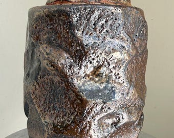 Pode incluir: Um pote cilíndrico de cerâmica com tampa, apresentando uma superfície rugosa e fortemente texturizada. O exterior exibe uma paleta de cores manchadas em tons de castanho escuro, bronze e preto com um brilho metálico e reentrâncias profundas.