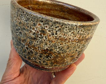 Pode incluir: Uma pequena tigela de cerâmica redonda com um esmalte texturizado e salpicado em tons de castanho, bege, cinzento e preto. O exterior apresenta uma superfície rugosa e manchada, enquanto o interior é liso com um acabamento castanho escuro.