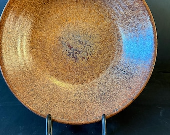 Pode incluir: Um prato redondo de cerâmica com um esmalte texturizado e salpicado em tons de laranja queimado, castanho escuro e cobre. A superfície apresenta um aspeto granulado com variações subtis de tom, criando um acabamento rústico e terroso.