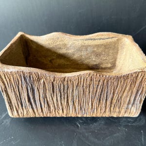 Pu&ograve; includere: Un vaso rettangolare in ceramica con un esterno strutturato simile alla corteccia in tonalit&agrave; marrone e marrone chiaro. Il contenitore presenta un bordo superiore ondulato e irregolare e un interno cavo, con un aspetto grezzo e organico.