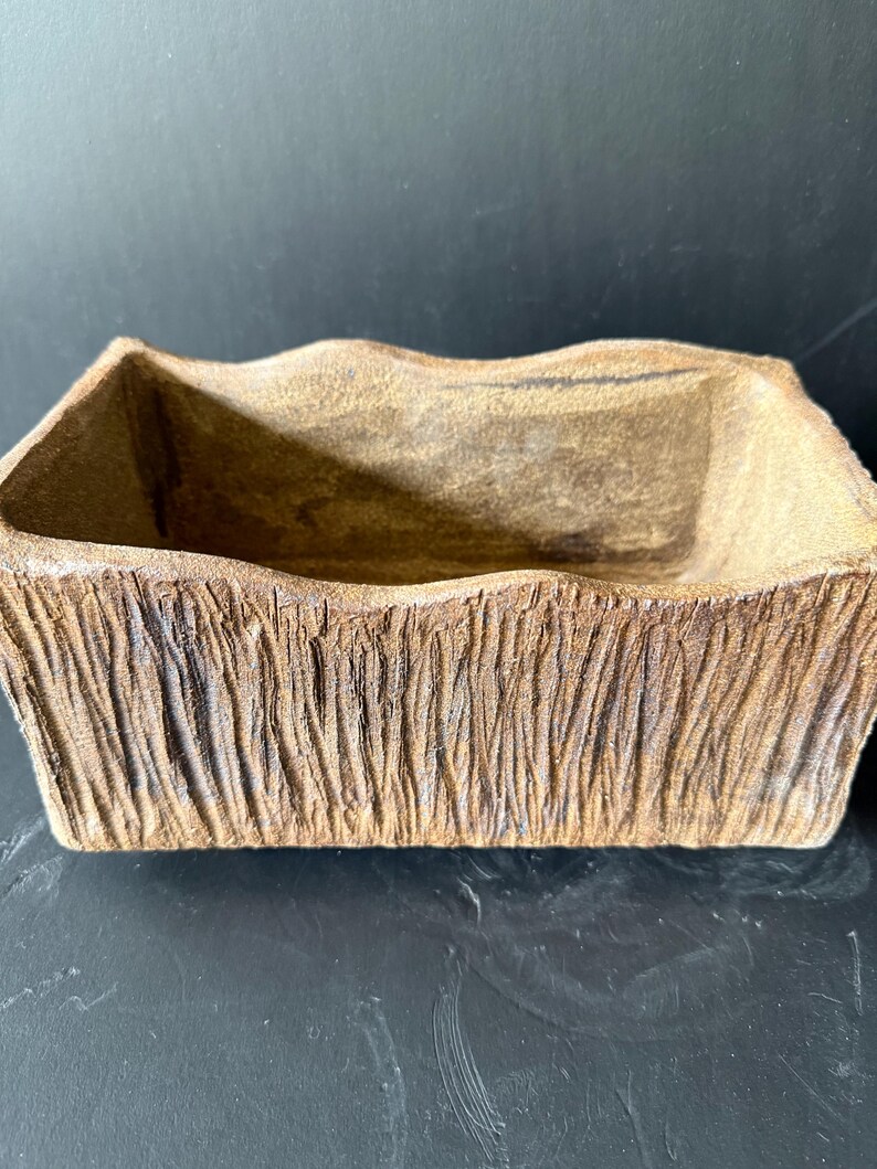 Pu&ograve; includere: Un vaso rettangolare in ceramica con un esterno strutturato simile alla corteccia in tonalit&agrave; marrone e marrone chiaro. Il contenitore presenta un bordo superiore ondulato e irregolare e un interno cavo, con un aspetto grezzo e organico.