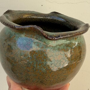 May include: A round ceramic pot with a wide, wavy rim. The glaze features a mottled blend of deep forest green, earthy brown, and hints of iridescent blue. The base is made of a textured, unglazed light beige clay, contrasting with the glossy finish.