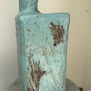 May include: A rectangular ceramic vase with a textured, light blue glaze and dark brown, rough patches. The vase features a narrow, cylindrical neck positioned on one side of the top edge, with a flat, squared-off body and a distinct, uneven surface.