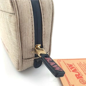 Pu&ograve; includere: Primo piano di una custodia in tela beige con una cerniera nera e un tirazip dorato. Il tirazip ha la parola "RAW" stampata in rosso. Un'etichetta di carta marrone con la parola "RAW" &egrave; visibile.