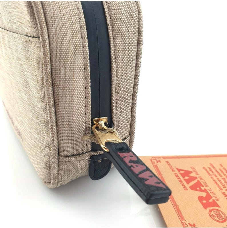 Pu&ograve; includere: Primo piano di una custodia in tela beige con una cerniera nera e un tirazip dorato. Il tirazip ha la parola "RAW" stampata in rosso. Un'etichetta di carta marrone con la parola "RAW" &egrave; visibile.