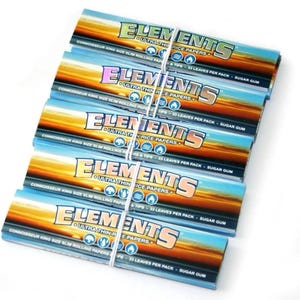 May include: A stack of Elements ultra-thin rice rolling paper packs. Each pack features a sunset design with the brand name in a bold, colorful font. The text includes "Connoisseur King Size Slim Rolling Papers & Tips - 33 Leaves Per Pack - Sugar Gum."