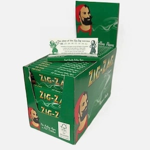 May include: A green cardboard display box containing multiple packs of Zig-Zag rolling papers. The box features the brand logo and a bearded man in a red hat. A single pack sits on top, displaying the text The story of the Zig-Zag success.