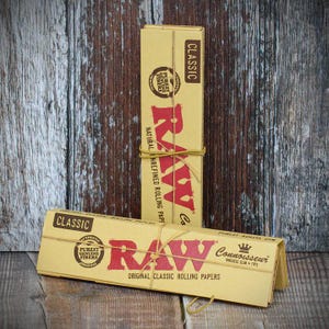 Puede incluir: Dos paquetes de papel de liar RAW Classic. Las cajas son de color beige con letras rojas y una banda marr&oacute;n. Una caja est&aacute; de pie y la otra est&aacute; tumbada. El texto de las cajas incluye "Classic", "RAW" y "Original Classic Rolling Papers".