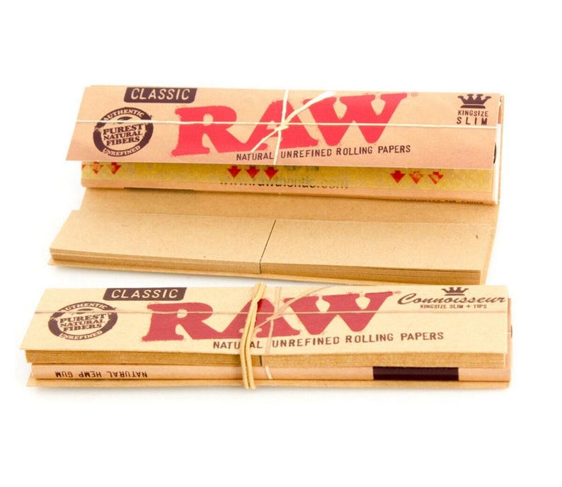 Puede incluir: Dos paquetes de papel de liar RAW Classic. El embalaje es de color beige con texto rojo que dice "RAW" y "NATURAL UNREFINED ROLLING PAPERS". La pila superior est&aacute; atada con hilo natural. El texto "CLASSIC" est&aacute; impreso en la parte superior.