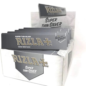 Puede incluir: Una caja expositora de papel de fumar Rizla+ Super Thin Silver. El embalaje es principalmente gris y blanco, con el nombre de la marca y los detalles del producto bien visibles. También se ve el texto "King Size Slim".