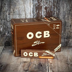 以下が含まれることがあります： OCB Slimのローリングペーパーの茶色の箱が2つ。大きな箱には白いOCBのロゴがあり、その下に「Slim」という文字があります。小さな箱にもOCBのロゴと「Slim」があり、「Virgin Paper」という文字があります。