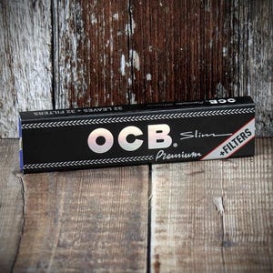 Può includere: Una scatola nera di cartine OCB Slim Premium con filtri. La scatola ha testo bianco e un logo OCB argentato. La scatola è su una superficie di legno.