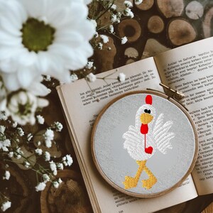 Könnte beinhalten: Gesticktes Hühnerdesign auf einem grauen Stoffreifen. Das Huhn ist weiß mit rotem Kamm und Kehllappen und gelben Beinen. Der Reifen liegt auf einem offenen Buch mit weißen Blumen im Hintergrund.