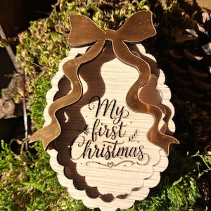 Peut inclure: Une décoration de Noël en bois de forme ovale avec un bord festonné, surmontée d'un nœud de couleur or métallique. Le centre est gravé du texte My first Christmas en écriture cursive, agrémenté d'un petit symbole de cœur.