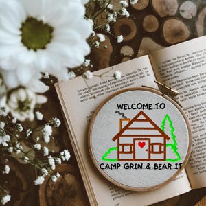 Könnte beinhalten: Ein handgestickter Stickrahmen mit einem Hüttendesign und dem Text "WELCOME TO CAMP GRIN & BEAR IT". Das Design umfasst ein rotes Herz, grüne Bäume und eine braune Hütte. Der Rahmen liegt auf einem offenen Buch.