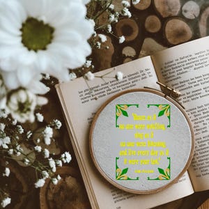 Könnte beinhalten: Ein bestickter Stickrahmen mit einem Zitat, der auf einem offenen Buch liegt. Das Zitat lautet: "Tanze, als würde niemand zusehen, singe, als würde niemand zuhören, und lebe jeden Tag, als wäre es dein letzter." Weiße Blumen und Holzscheiben im Hintergrund.