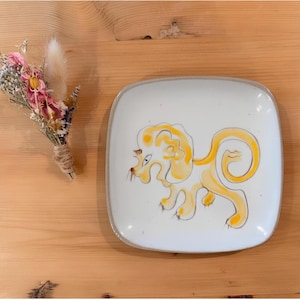 May include: A square white ceramic plate featuring a hand-painted yellow lion illustration with dark outlines, accompanied by a small bouquet of dried flowers in shades of pink, white, and green, tied together with natural twine.