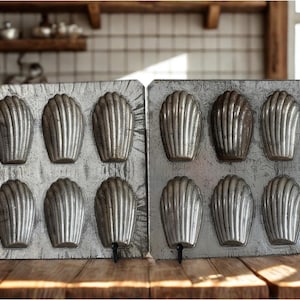 Könnte beinhalten: Zwei rechteckige, vintage Madeleine-Backformen. Jede Form hat sechs muschelförmige Vertiefungen. Die Formen bestehen aus einem matten, silberfarbenen Metall und stehen auf einer Holzoberfläche. Der Hintergrund zeigt ein Regal mit Küchenutensilien.