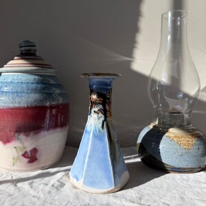 May include: Three ceramic pieces: a lidded jar with blue, red, and white glaze, a blue vase with a geometric shape, and a glass oil lamp with a ceramic base. The jar has a floral design. The lamp has a clear glass chimney.