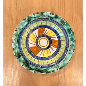 May include: A round ceramic plate featuring a scalloped edge with a mottled green glaze. The center displays a radial pattern of yellow and orange segments surrounded by concentric rings in shades of blue, light green, and dark green.