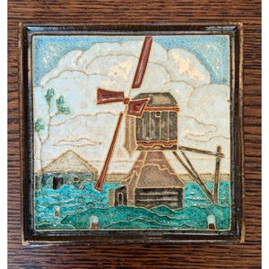 May include: A square ceramic tile featuring a relief design of a windmill in brown and dark tones set against a light blue and white sky. The foreground displays textured green landscape elements and a small structure, all framed by a dark border.