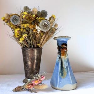 May include: A metallic, tarnished silver-toned vase filled with a bouquet of dried yellow flowers, wheat, and grey globe thistles, alongside a small, hexagonal blue ceramic vase with brown glaze accents and a small bundle of dried pink flowers.