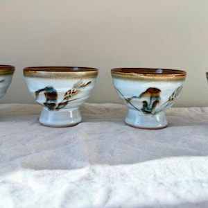 Puede incluir: Cuatro cuencos de cerámica con base blanca, borde marrón y diseños florales decorativos marrones y azules. Los cuencos tienen una base de pedestal y están dispuestos en fila sobre un paño blanco.