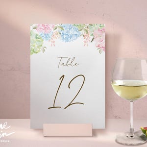 May include: A white table number card with floral accents in pink, blue, and green. The card displays the word "Table" and the number "12" in a brown script font. A wine glass with white wine and figs are in the foreground.