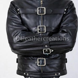 Könnte beinhalten: Schwarze Lederjacke mit mehreren Riemen und silbernen Schnallen. Die Jacke hat einen hohen Kragen und einen Reißverschluss. Der untere Teil der Jacke hat einen Schrittriemen mit Schnalle und Metallring. Der Text "HMleathercreations" ist sichtbar.
