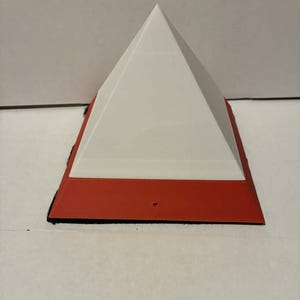 May include: A white pyramid-shaped object sits on a red rectangular base with a black edge. The pyramid has a smooth surface and a sharp point. The base is flat and appears rigid. The background is a plain white surface.