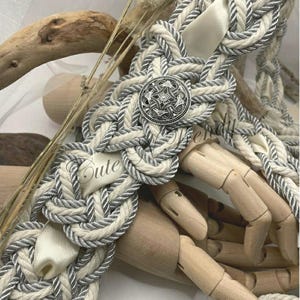 May include: A decorative rope belt with a silver button and the word "Cute" on a white ribbon. The belt is made of cream-colored rope with gray accents and a cream-colored satin ribbon.